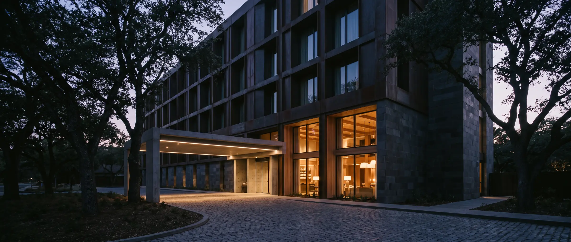 Boutique hotel exterior at dusk with a warmly lit double-height lobby and a porte-cochere