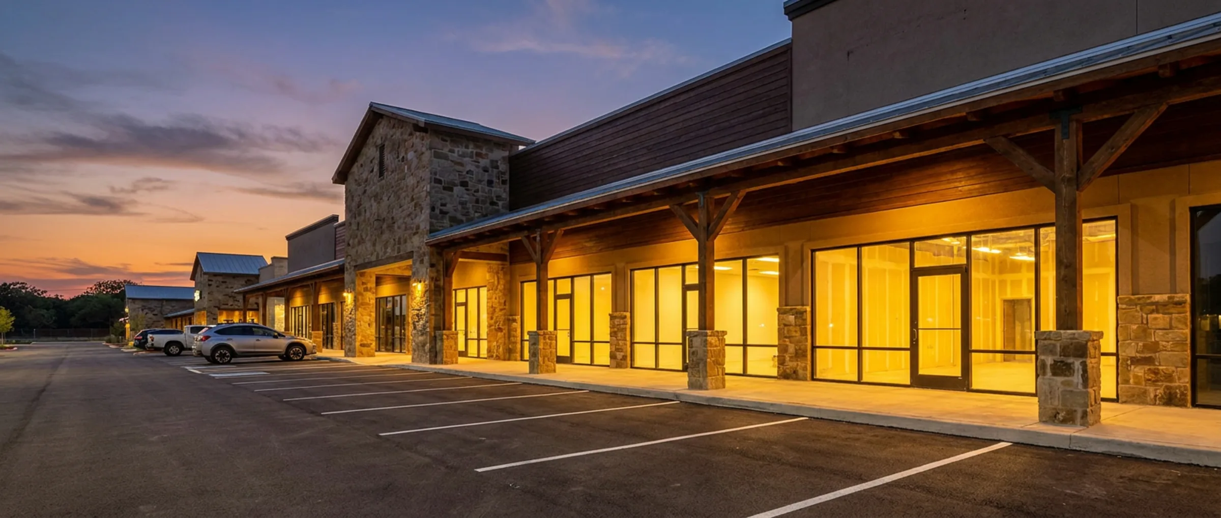 Neighbourhood retail centre at dusk with a stone-columned covered walkway and warmly lit storefronts