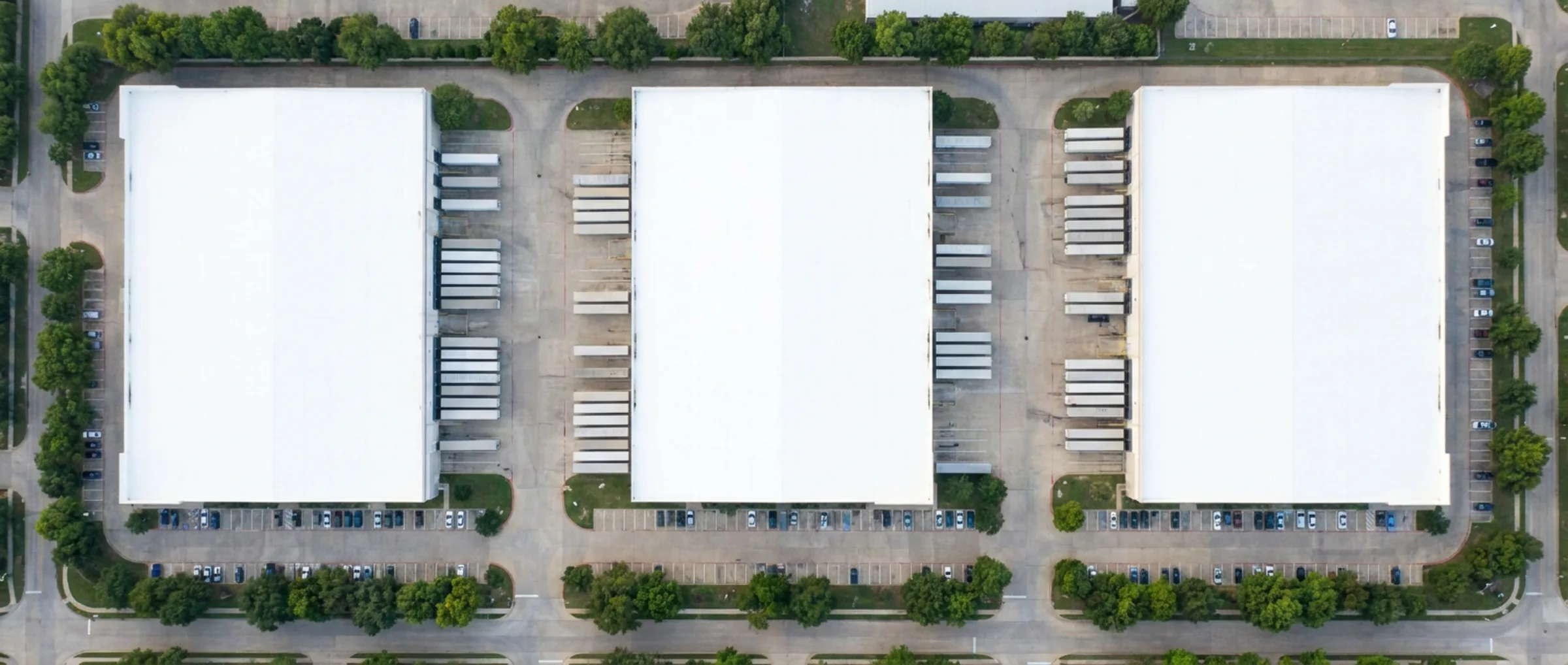 Aerial view looking down at three adjacent warehouse buildings with loading aprons, parked trailers, and tree-lined streets around them