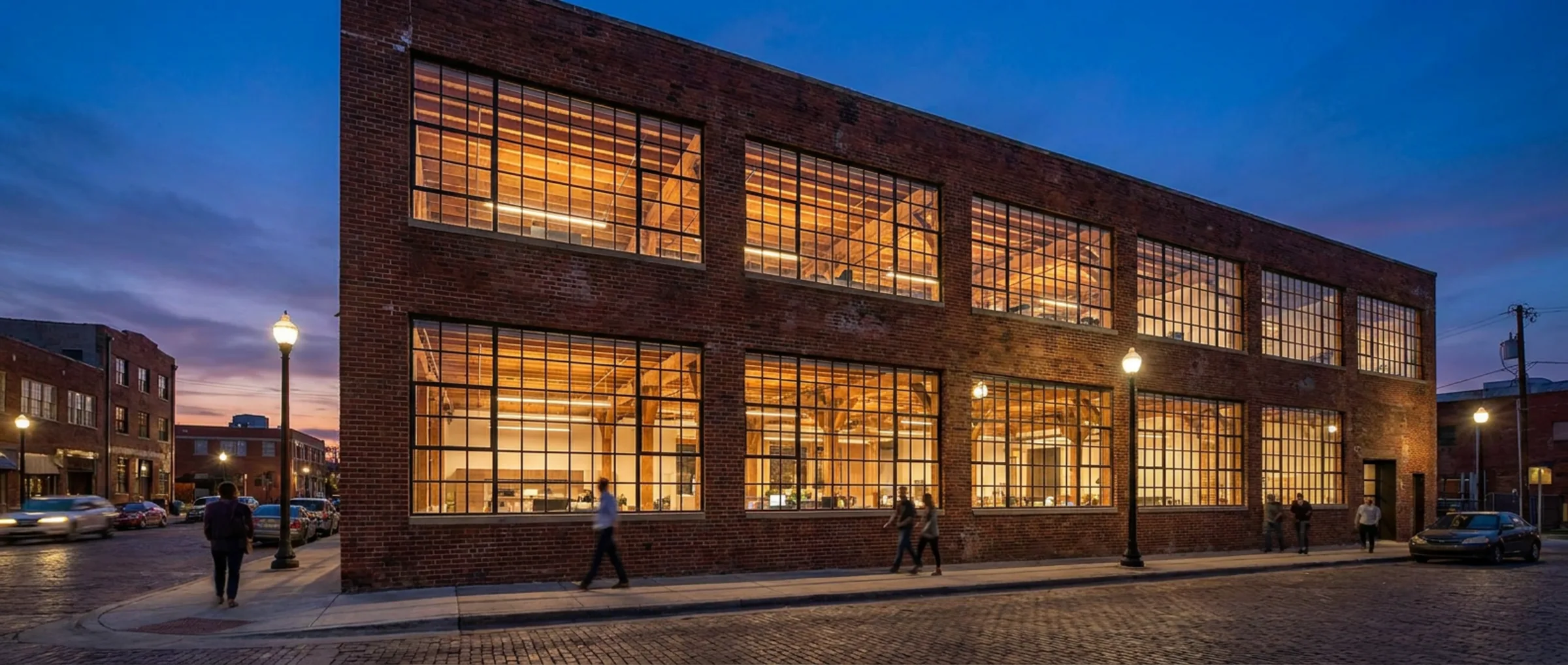 Restored red brick warehouse office at dusk, with tall steel-framed windows and warm interior light on an urban street